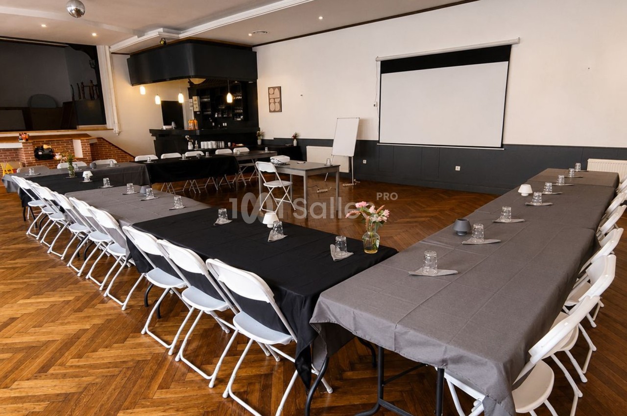 Salle de réunion avec tables disposées en U, chaises pliantes, écran de projection et parquet en bois.