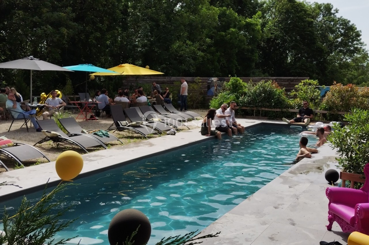 Piscine extérieure entourée de chaises longues, avec des personnes se relaxant et discutant sous des parasols colorés.