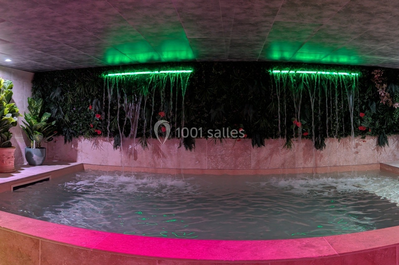 Piscine intérieure avec cascades murales, éclairage coloré et décor végétal luxuriant.
