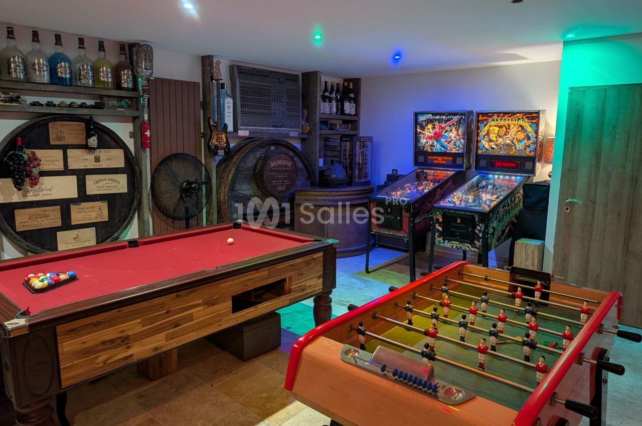 Salle de jeux avec billard, baby-foot, flippers et décorations de bar incluant tonneaux et bouteilles.