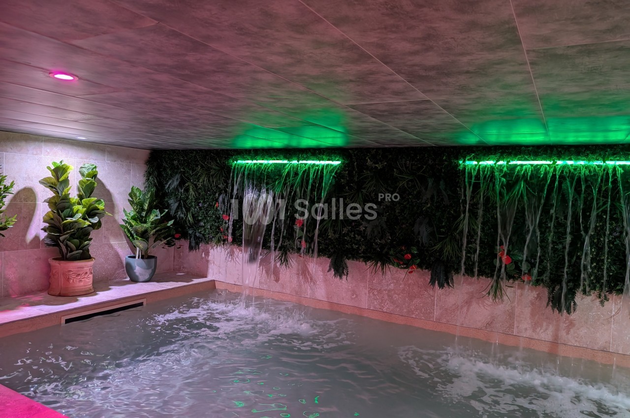 Piscine intérieure avec murs végétalisés, cascades d'eau et éclairage tamisé rose et vert.