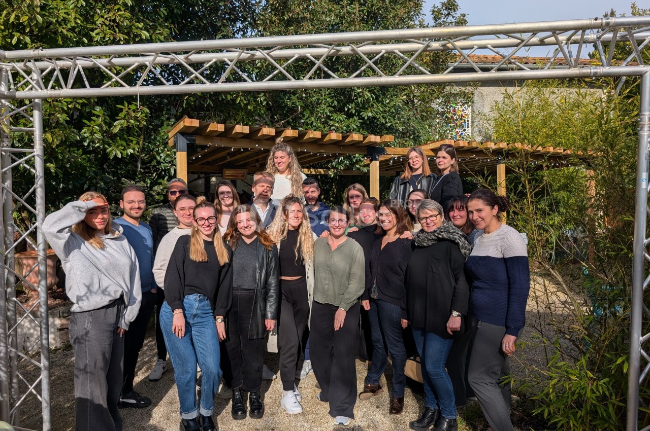 Groupe de personnes posant ensemble à l'extérieur, devant une pergola et des arbres, par une journée ensoleillée.