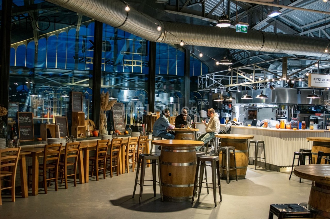 Intérieur d'un restaurant avec tables en bois, bar central éclairé et grandes baies vitrées donnant sur l'extérieur.