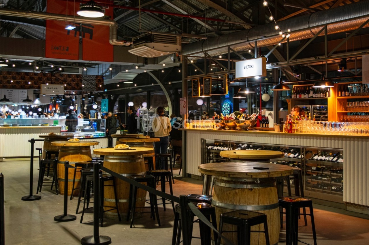 Intérieur d'un marché couvert avec un bar en bois, des tables rondes et des étals éclairés en arrière-plan.
