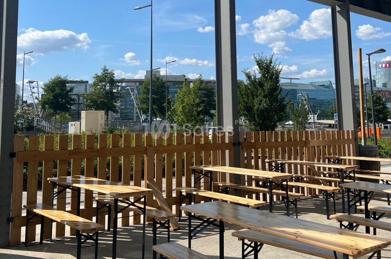 Terrasse extérieure avec tables et bancs en bois, entourée d'une clôture, vue sur des bâtiments modernes et un ciel dégagé.