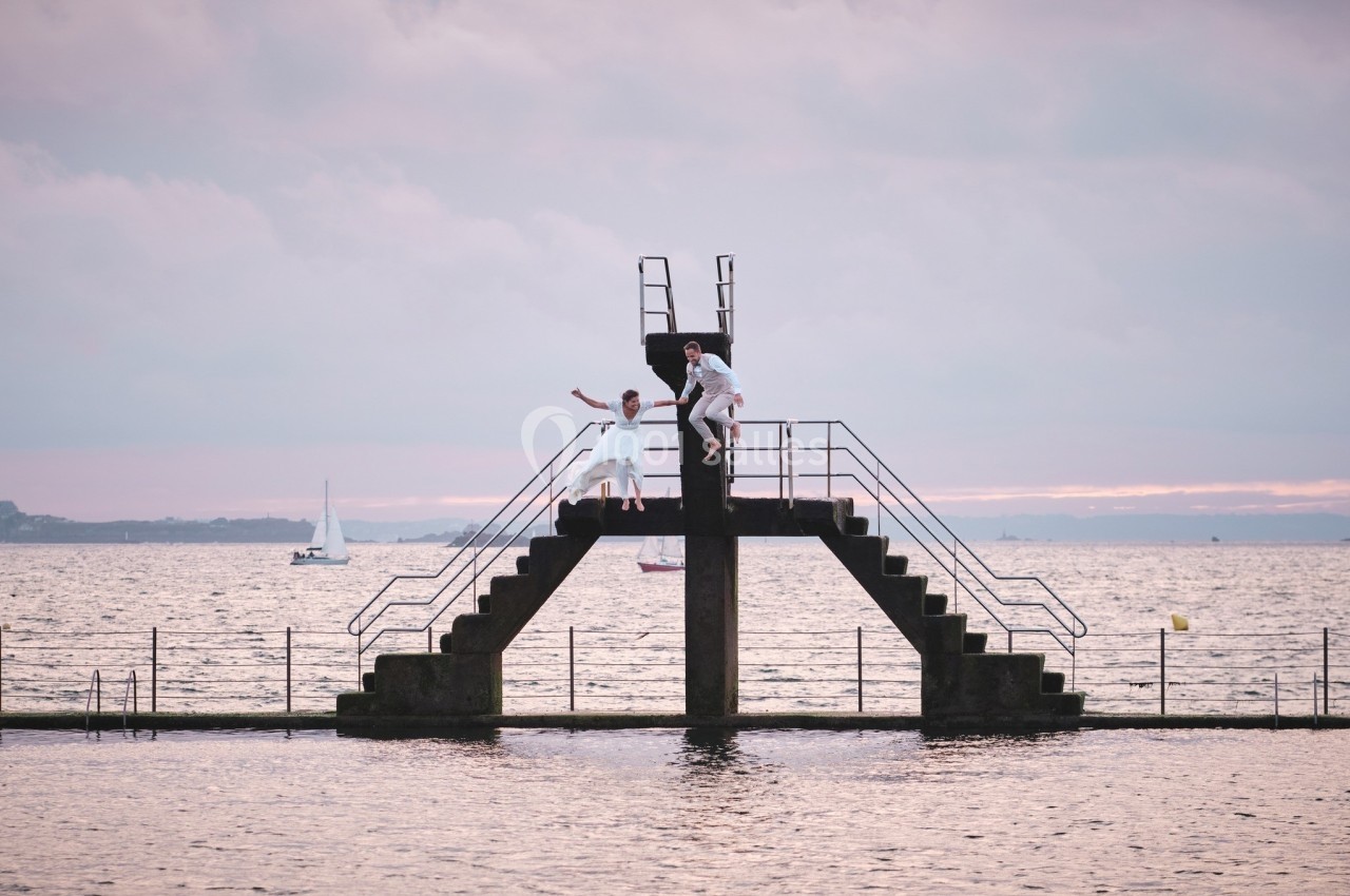 Deux personnes en blanc sur une plateforme de plongeon au bord de la mer, avec un ciel nuageux au coucher du soleil.