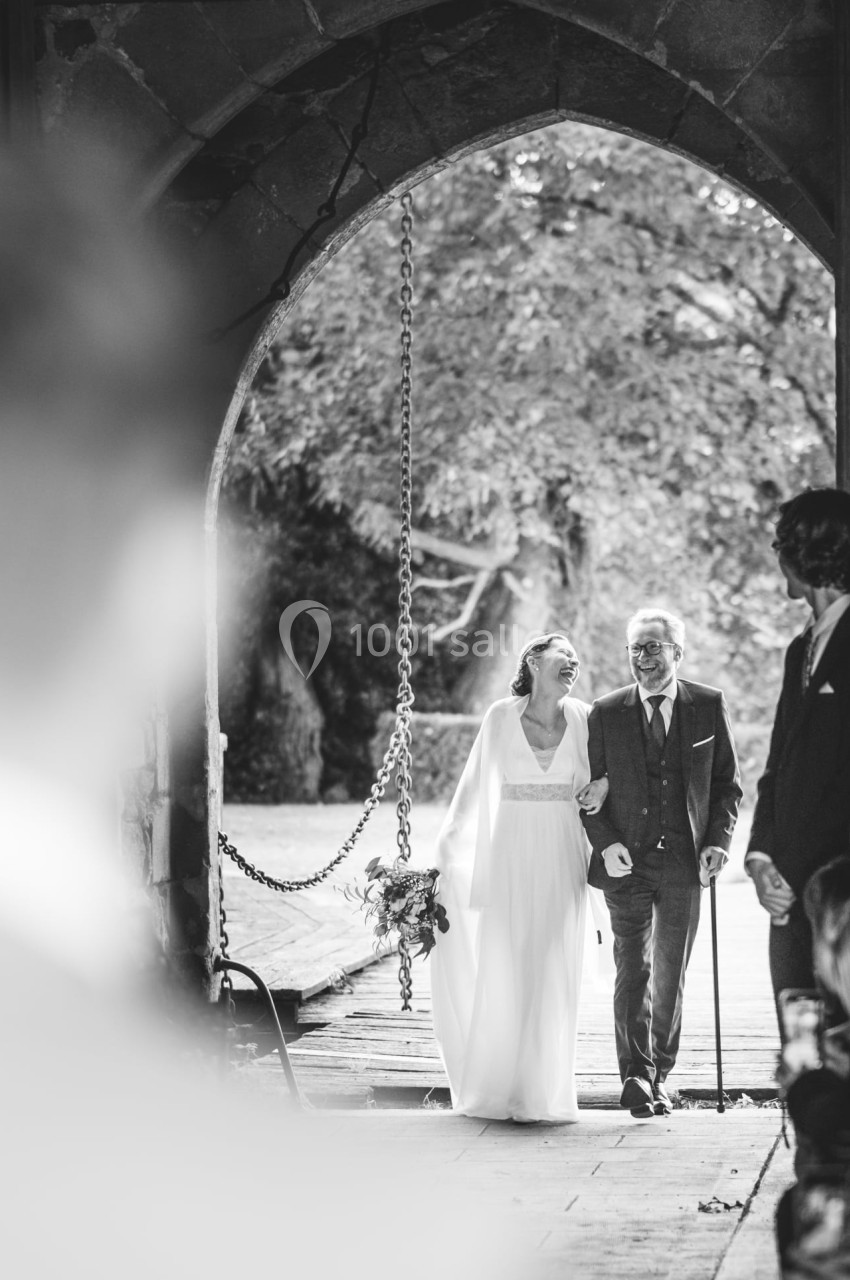 Une mariée souriante entre au bras d'un homme âgé sous une arche en pierre, entourée d'invités.