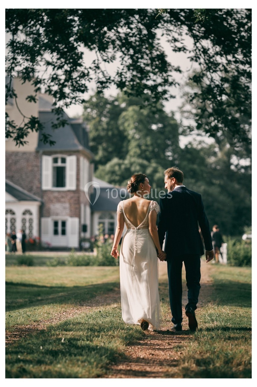Un couple en tenue de mariage marche sur un chemin de terre devant une maison entourée de verdure.