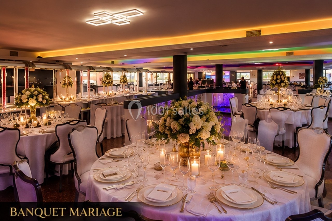 Salle de réception élégamment décorée pour un mariage, avec tables rondes, nappes blanches et arrangements floraux.