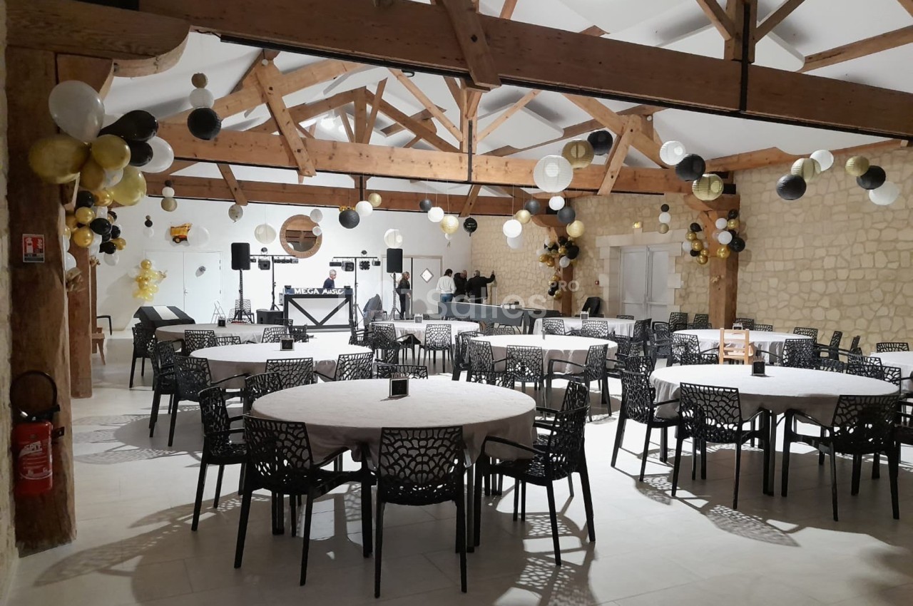 Salle de réception décorée avec des ballons noirs, blancs et dorés, tables rondes dressées et espace DJ au fond.