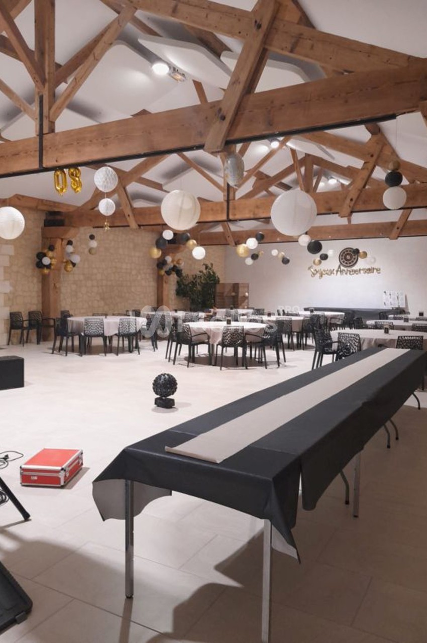 Salle de réception décorée pour un anniversaire, avec tables alignées, guirlandes et lanternes suspendues au plafond en bois.
