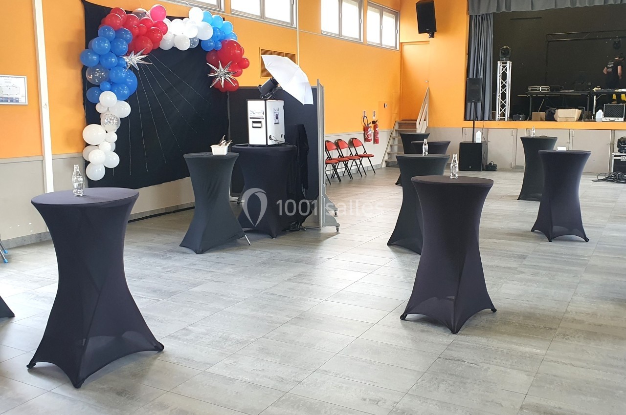 Salle décorée avec des ballons, tables hautes noires et un photobooth, prête pour un événement festif.