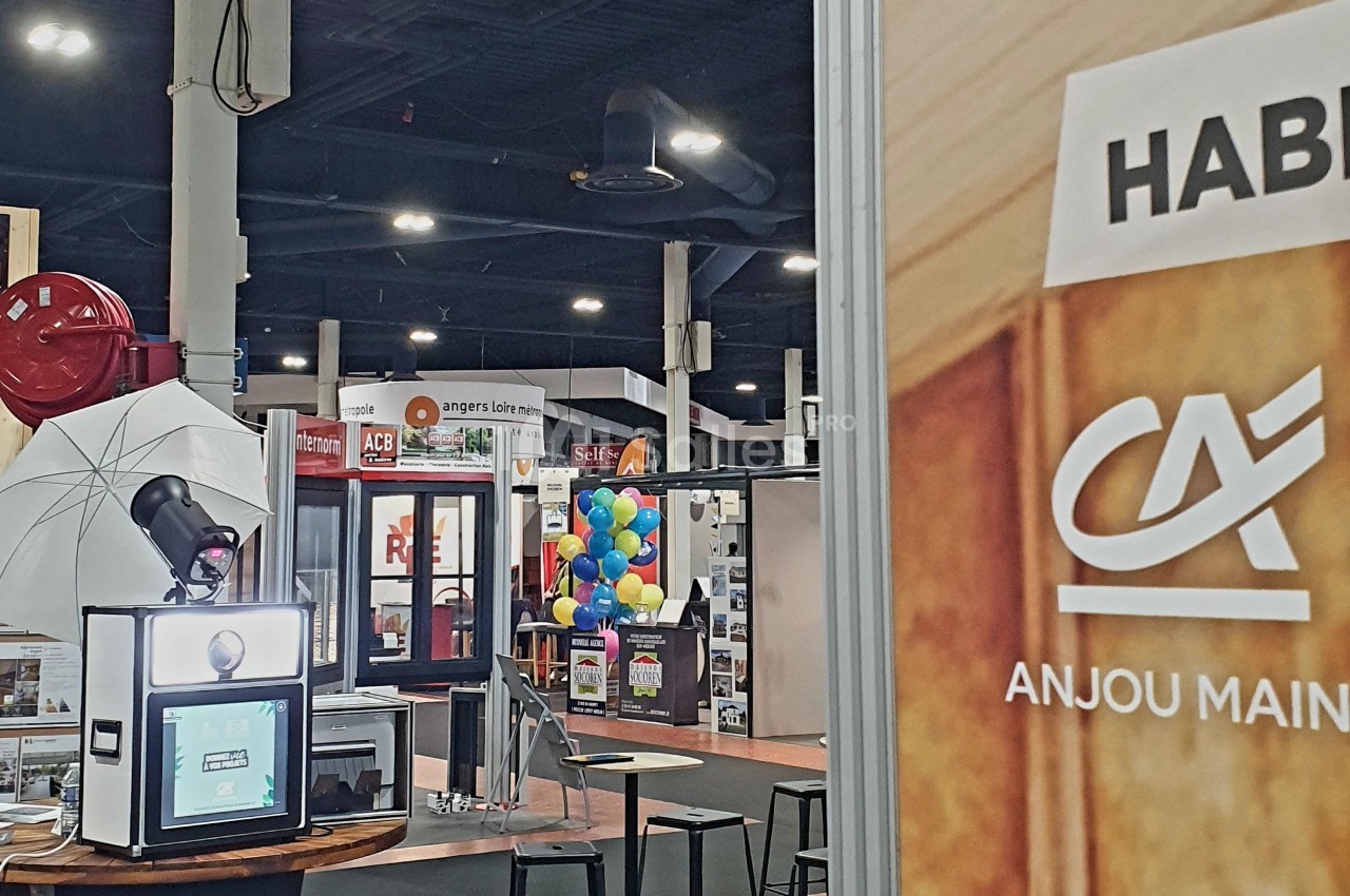 Borne photo avec éclairage et décor, installée dans un salon professionnel avec stands et visiteurs en arrière-plan.