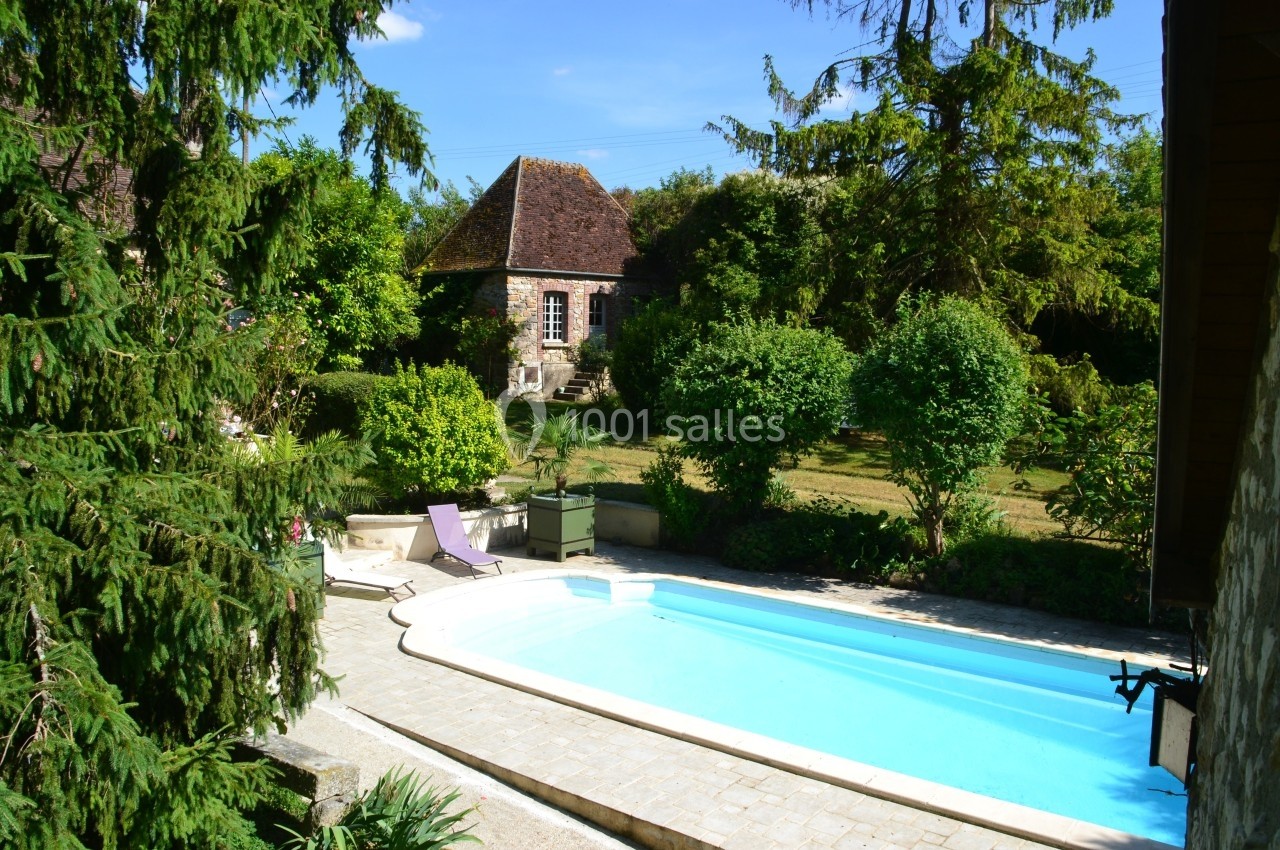 Piscine extérieure entourée de végétation et d'arbres, avec une maison en pierre au toit en pente en arrière-plan.