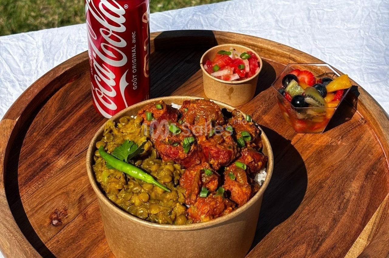 Repas servi sur un plateau en bois avec un bol de curry, des lentilles, une salade de fruits et une canette de soda.
