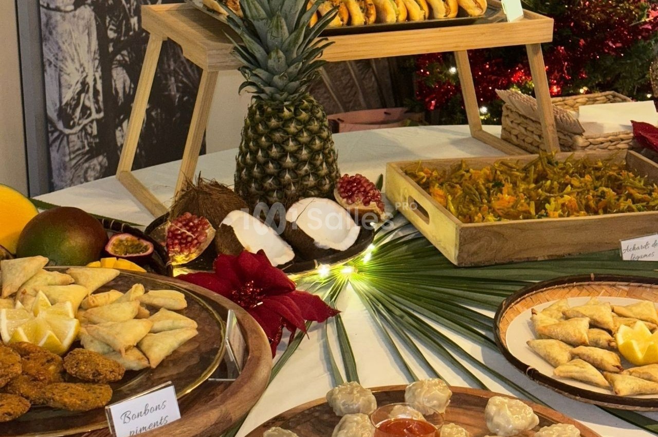 Buffet tropical avec fruits exotiques, samoussas, bouchées apéritives et décorations végétales sur une table blanche.