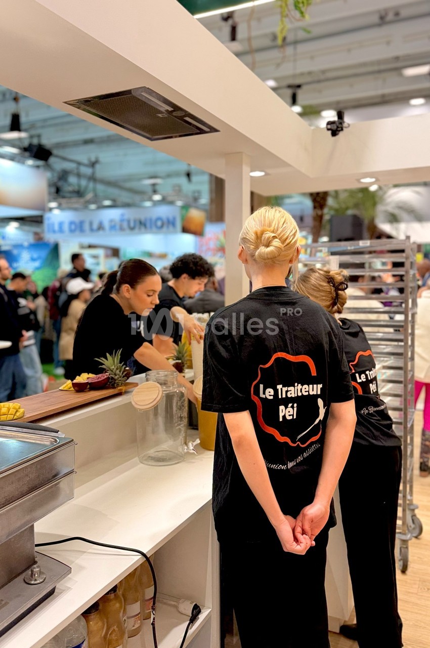 Des personnes préparent des plats dans un stand culinaire, avec des ingrédients et équipements visibles en arrière-plan.