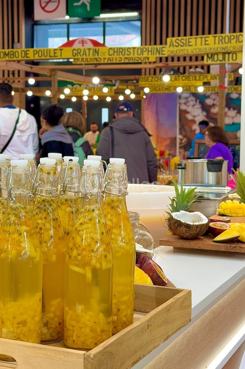 Bouteilles de jus tropical avec morceaux de fruits en avant-plan, stand de cuisine créole animé en arrière-plan.