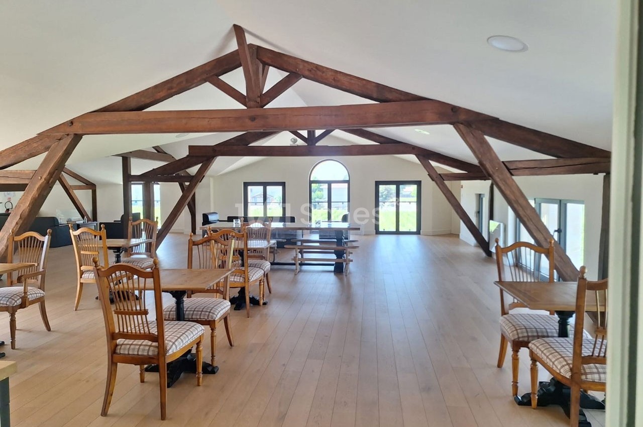 Salle lumineuse avec charpente en bois apparente, tables et chaises disposées sur un sol en parquet clair.