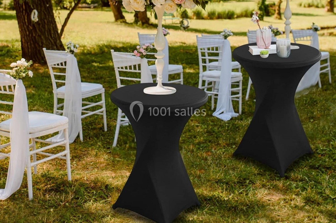 Tables hautes noires avec nappes, entourées de chaises blanches décorées, disposées dans un jardin pour un événement.