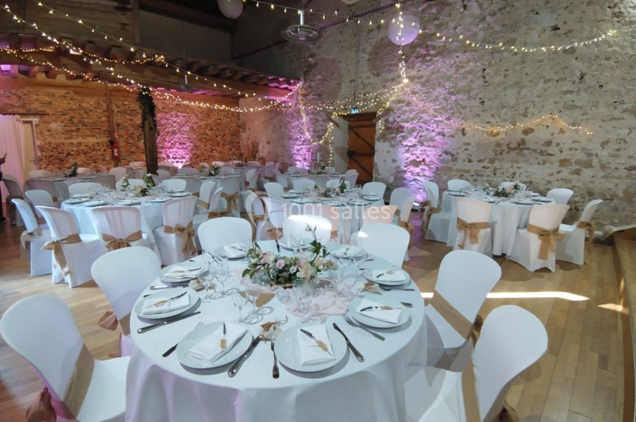 Salle de réception décorée pour un événement, avec tables rondes dressées, nappes blanches et guirlandes lumineuses.