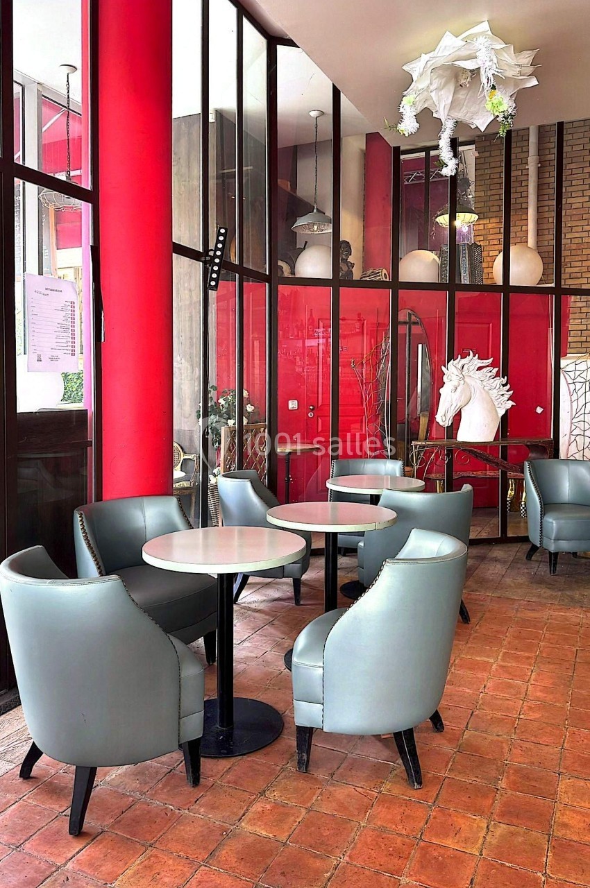 Salle lumineuse avec fauteuils gris autour de tables rondes, murs rouges et décoration moderne, dont une tête de cheval…