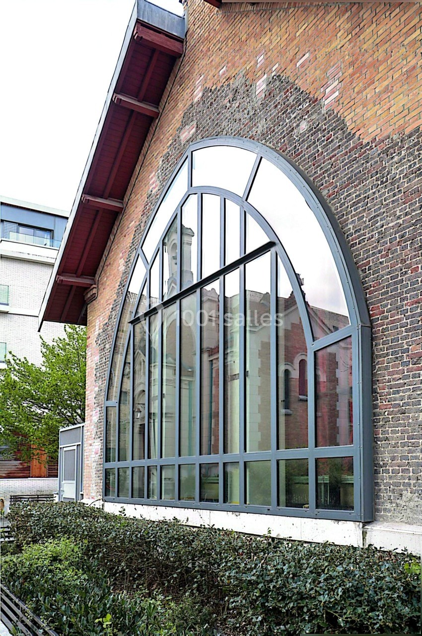 Grande baie vitrée en arc sur une façade en briques, reflétant partiellement un bâtiment voisin.
