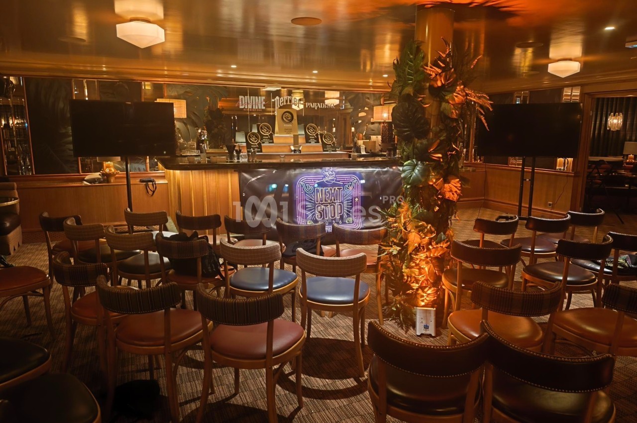 Salle aménagée avec des chaises disposées face à un bar éclairé, décoré d'un panneau lumineux et de plantes.