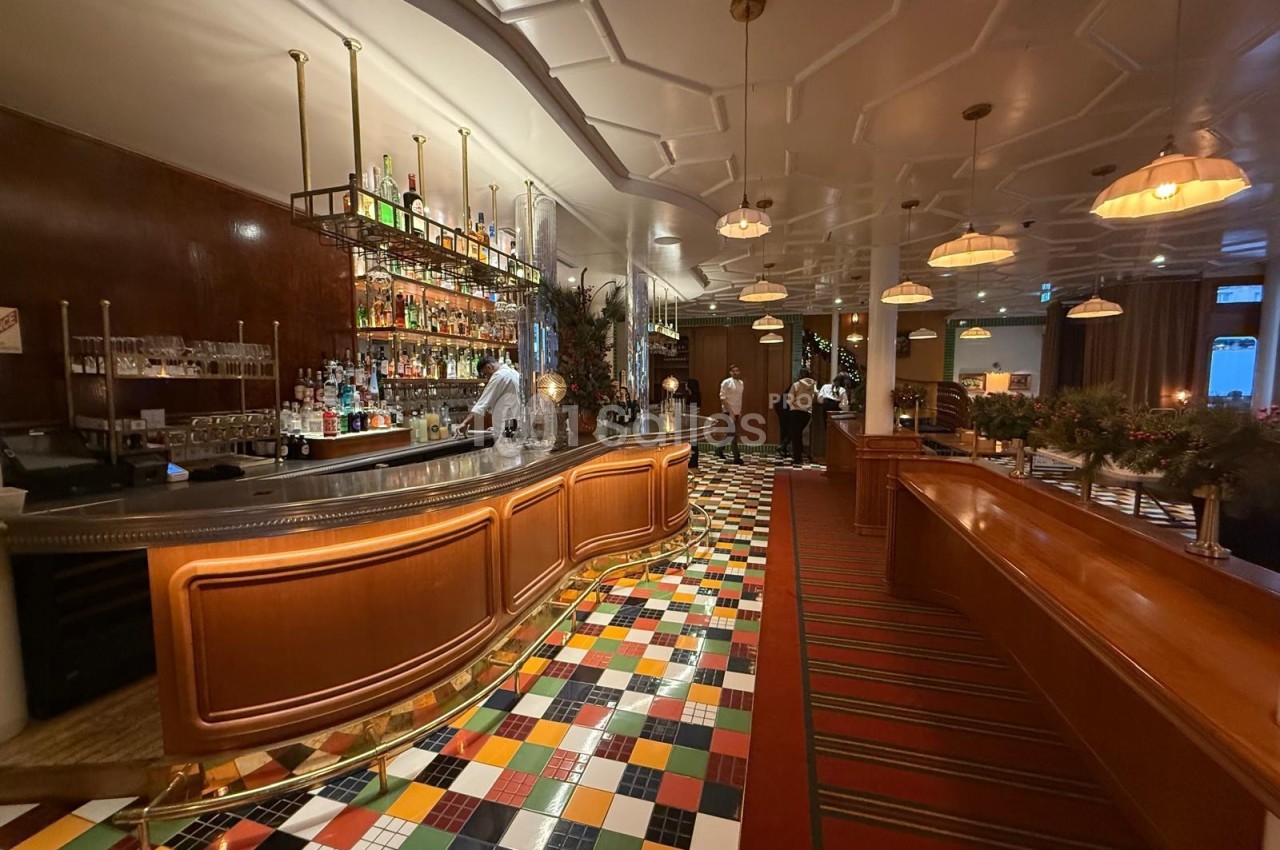 Intérieur d'un bar avec comptoir en bois, étagères de bouteilles, sol coloré et éclairage chaleureux.