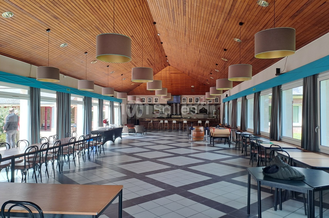 Salle spacieuse avec plafond en bois, grandes baies vitrées, tables et chaises disposées dans un espace lumineux.