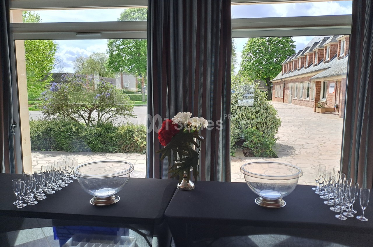 Table dressée avec des coupes et des bols en verre devant de grandes baies vitrées donnant sur un jardin et une cour pavée.