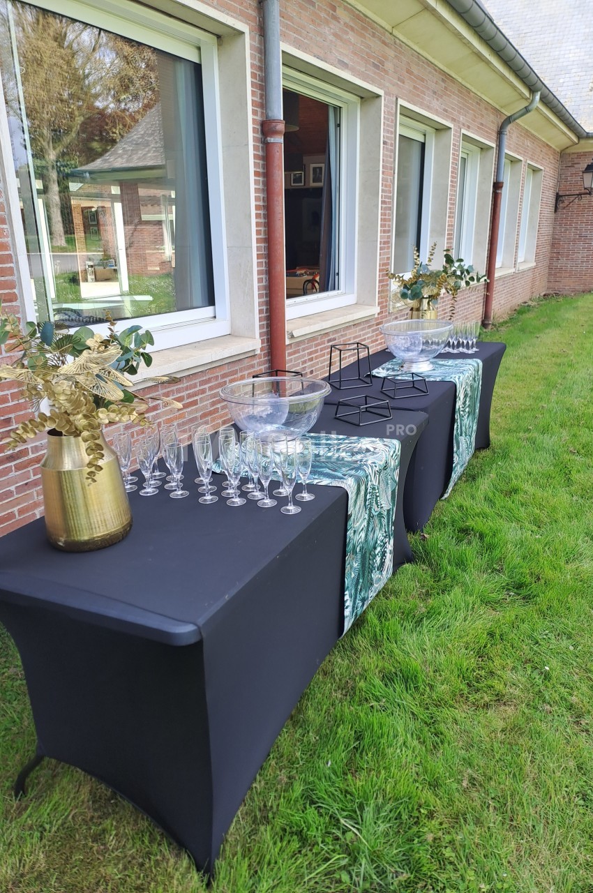 Tables noires dressées en extérieur avec verres, carafes et décorations florales devant un bâtiment en briques.
