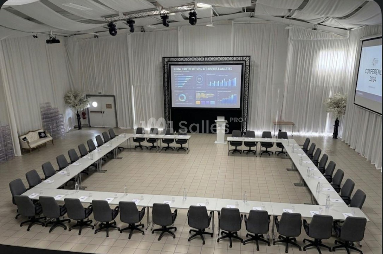 Salle de conférence avec disposition en U, tables blanches, chaises grises et écrans affichant des graphiques.