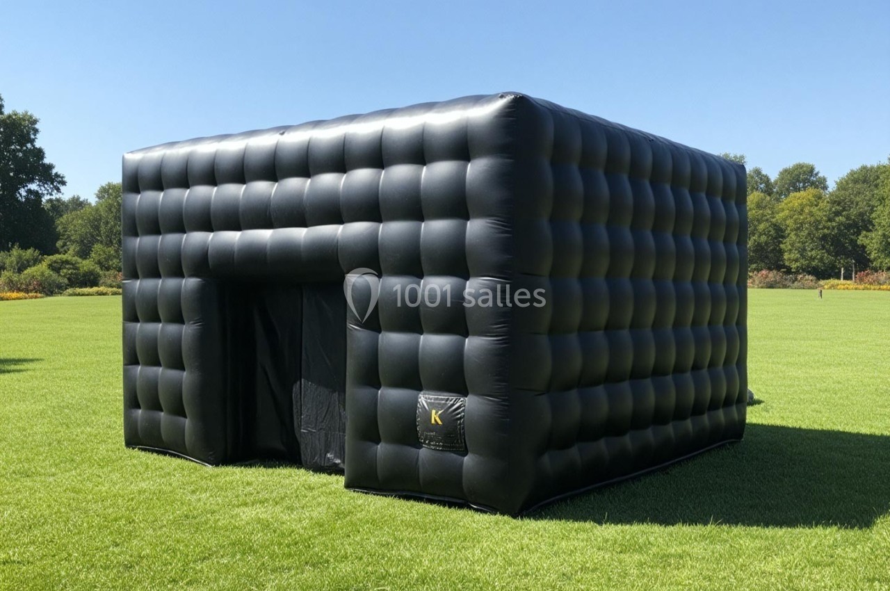 Structure gonflable noire en forme de cube, installée sur une pelouse verte sous un ciel dégagé.