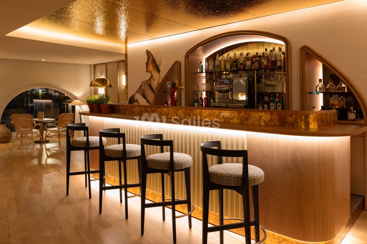 Bar intérieur avec comptoir courbe éclairé, tabourets noirs, étagères de bouteilles et mobilier lounge en arrière-plan.