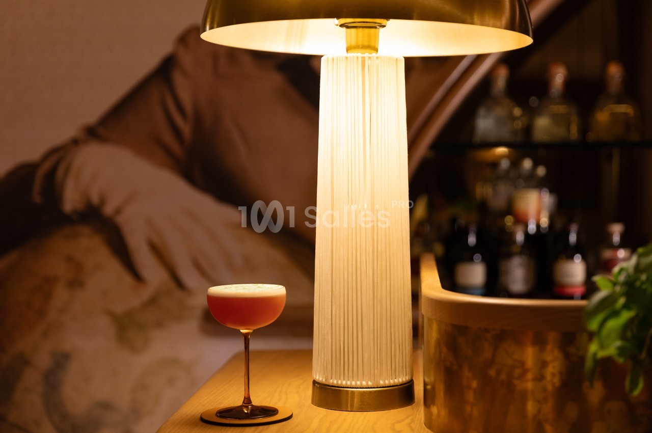 Lampadaire design en laiton avec abat-jour plissé blanc éclairant un verre de cocktail rouge sur comptoir.