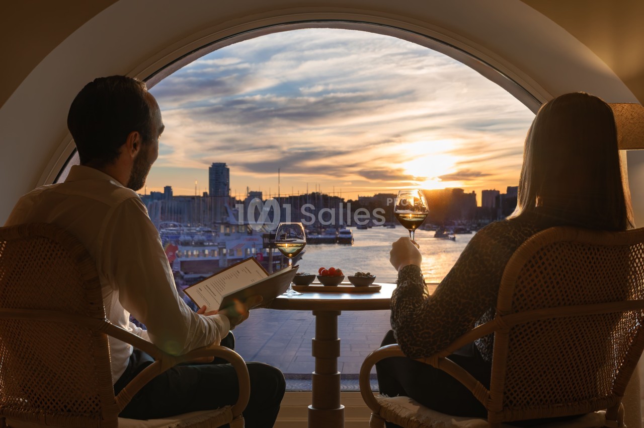 Deux personnes assises face à une baie vitrée panoramique surplombant le Vieux-Port au coucher du soleil, verres de vin blanc à la main.