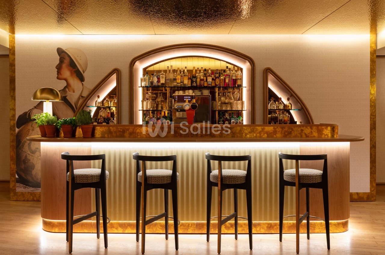 Bar avec comptoir en bois clair, quatre tabourets noirs, étagères illuminées de bouteilles et décoration art déco en relief.