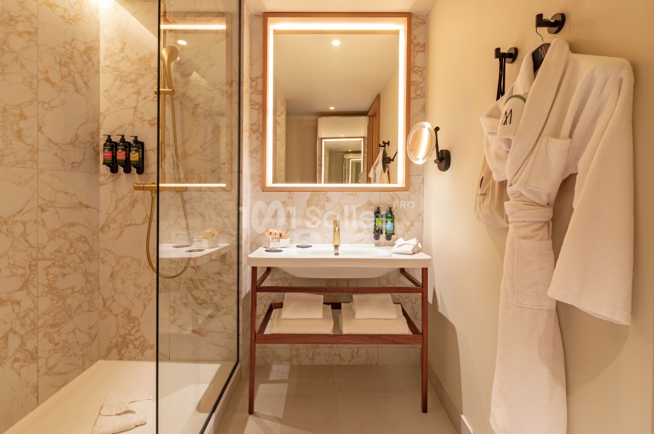 Salle de bain carrelée de marbre clair avec douche vitrée, vasque et miroir rétroéclairé, serviettes et produits de toilette.