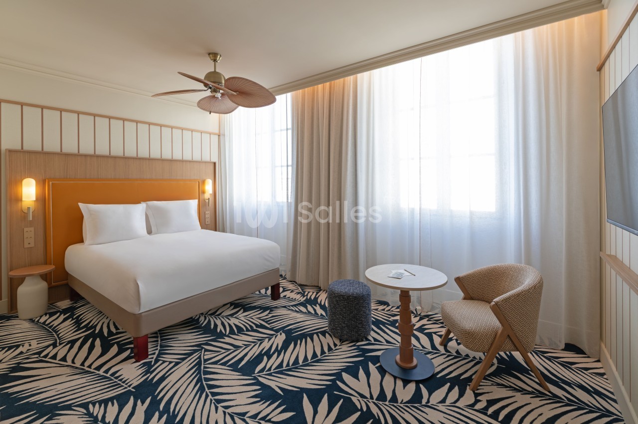 Chambre d'hôtel avec lit double aux draps blancs, fauteuil beige, petite table ronde et larges baies vitrées avec rideaux beige.