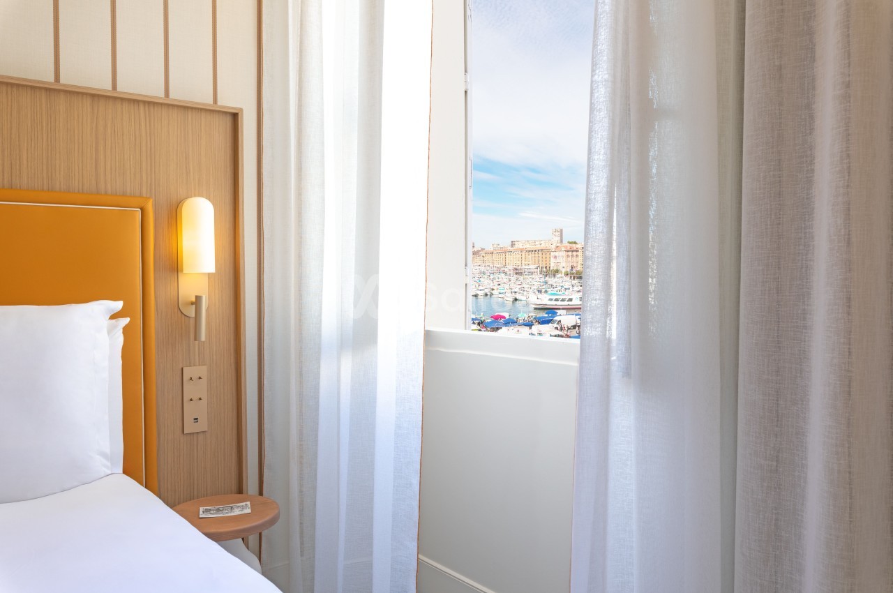 Chambre avec lit à tête de lit orange, vue sur le Vieux-Port de Marseille et ses bateaux.