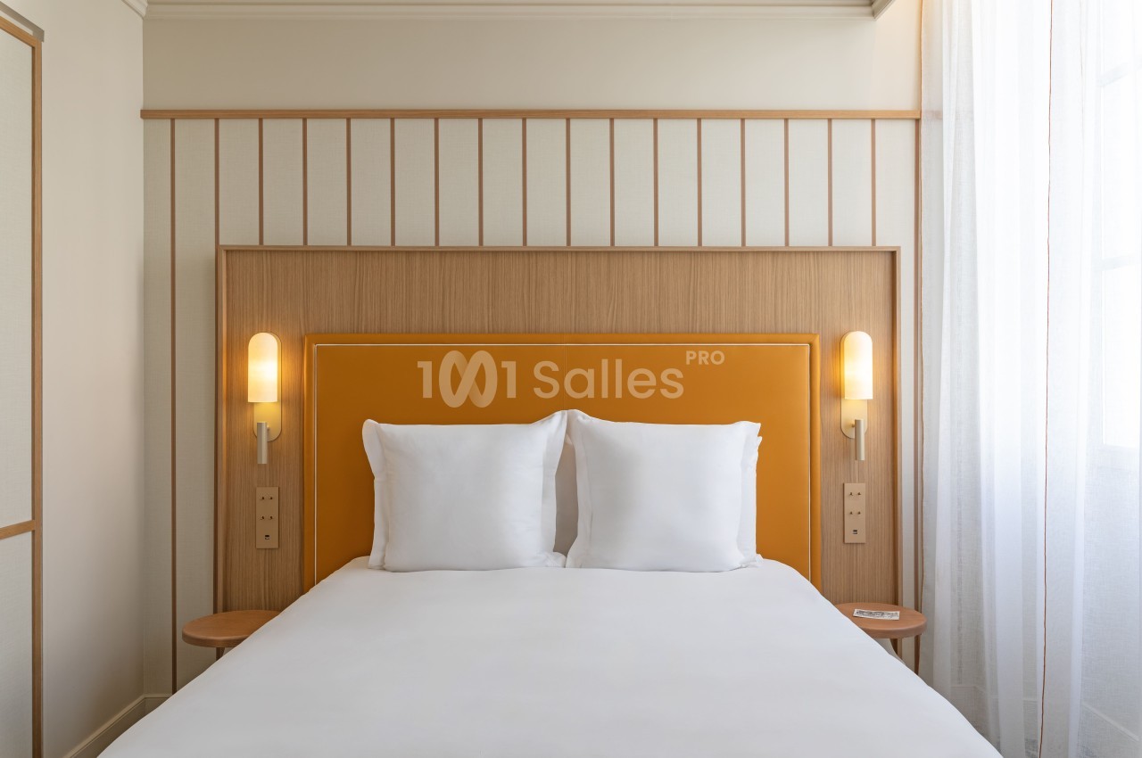 Chambre avec lit double, tête de lit en bois clair, deux lampes murales et linge blanc.