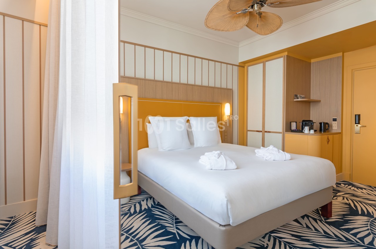 Chambre d'hôtel avec lit queen-size, tête de lit jaune moutarde, mobilier en bois clair et tapis à motifs géométriques.
