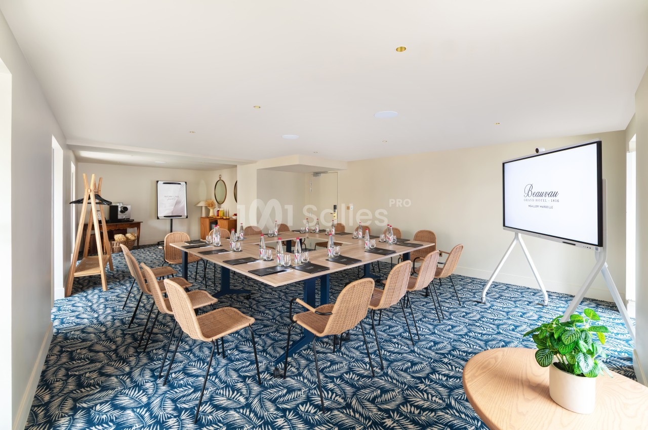 Salle de réunion dressée avec table rectangulaire, chaises en rotin, écran de projection, sol à motifs bleus et blancs.