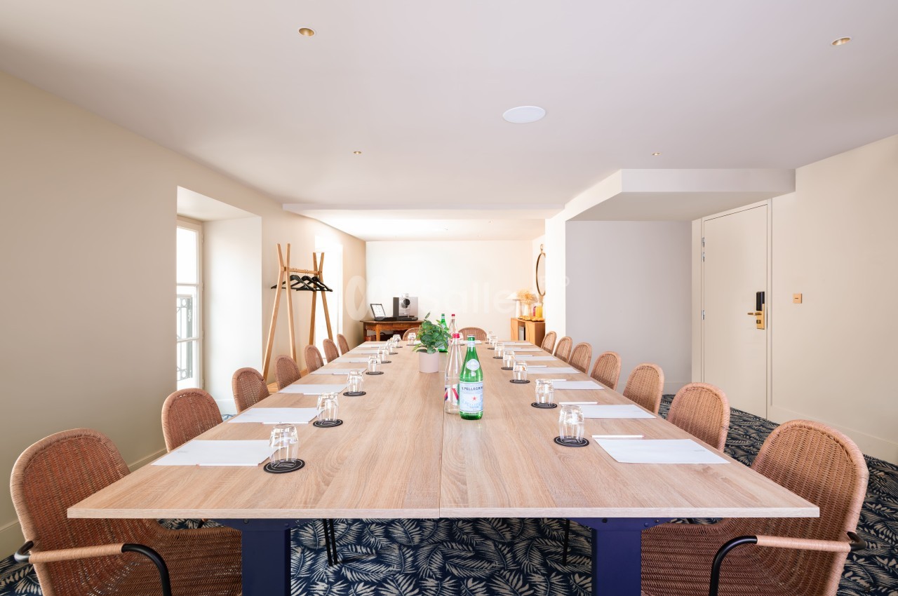 Salle de réunion avec table rectangulaire en bois clair, chaises en rotin, verres et carafes d'eau alignés.