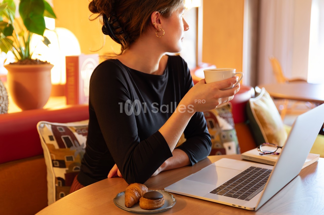 Femme assise à un bureau avec laptop, tenant une tasse de café, croissant et pâtisserie sur une assiette - intérieur lumineux aux murs jaunes.