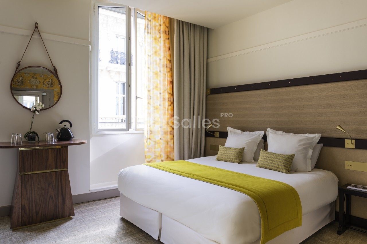 Chambre avec lit double blanc, linge de lit anis, fenêtre ouvrant sur la ville et mobilier bois.
