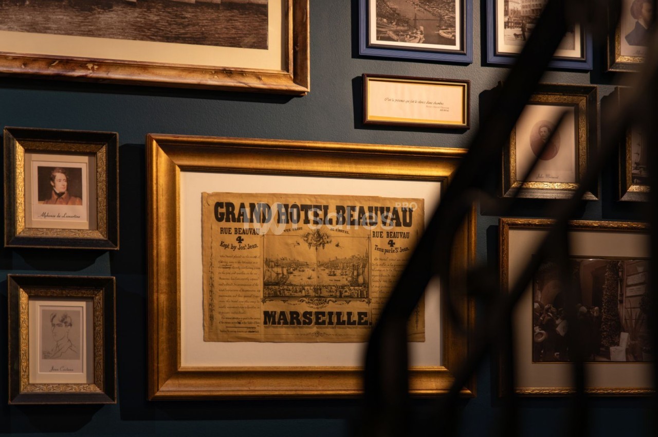 Mur bleu foncé orné de cadres dorés avec affiche historique du Grand Hôtel Beauvau et portraits anciens.
