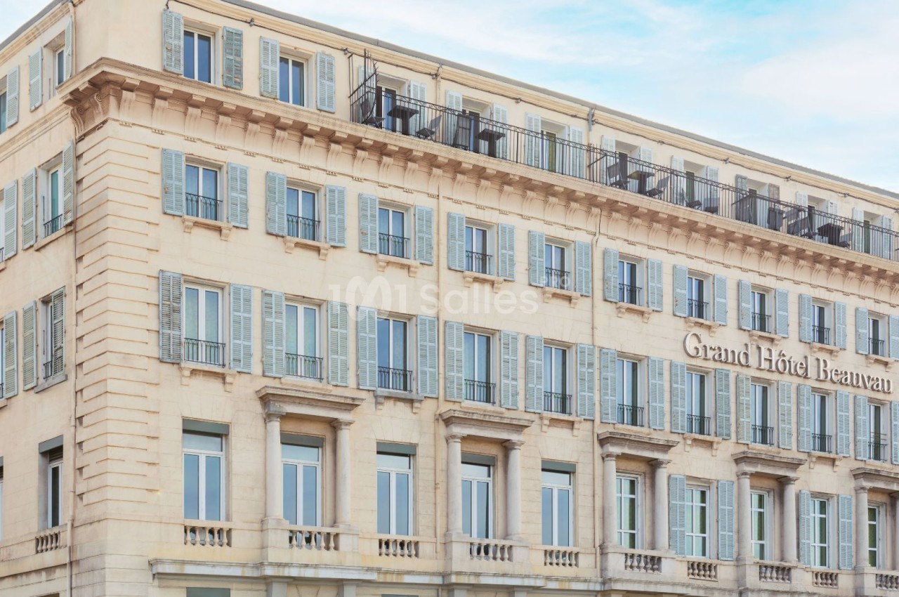 Immeuble haussmannien beige aux cinq étages avec volets bleus, balcons en fer forgé et balustrades ornementales.