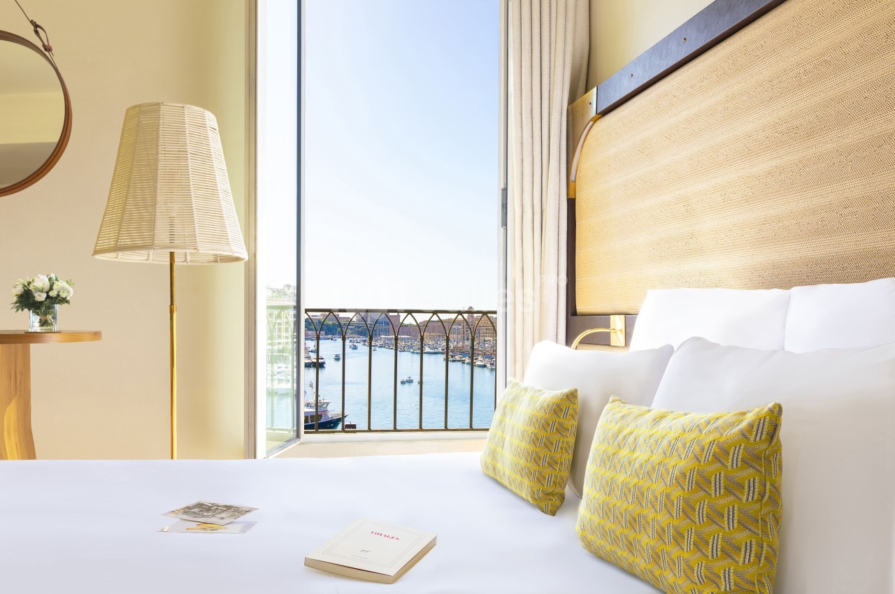 Chambre d'hôtel avec lit double, coussins jaunes, lampadaire, vue sur le Vieux-Port de Marseille par baie vitrée.